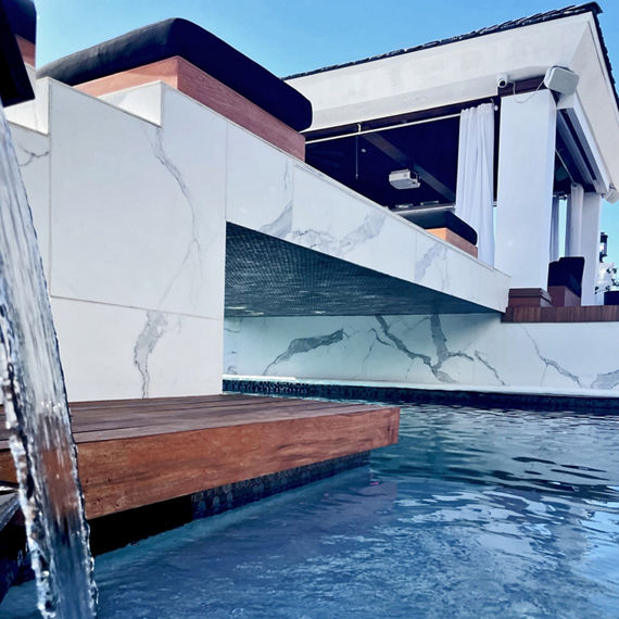 Jason Derulos backyard with pool deck and bridge made of black hexagon glass mosaic and black white and gray porcelain tile that looks like marble, and wood deck with lounge chairs.