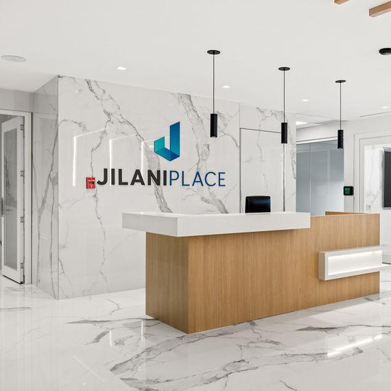 Welcome area with white marble look porcelain slab on floors and walls, light wood welcome desk, and office to right.