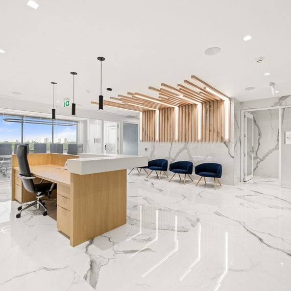 Welcome area with white marble look porcelain slab on floors and walls, light wood welcome desk, and three blue chairs.