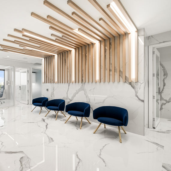 Welcome area with white marble look porcelain slab on floors and walls, light wood welcome desk, and three blue chairs.
