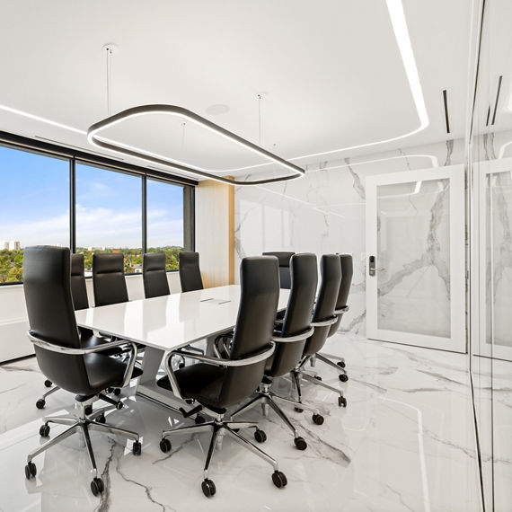 Work room with white marble look porcelain slab on back wall, black and white carpet, large windows showing outside view, and large white table with black sitting chairs.