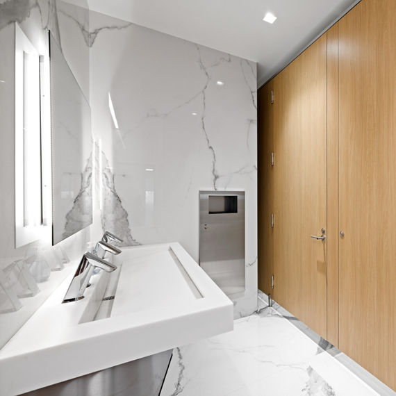 Bathroom with white marble look porcelain slab on walls and floor, light wood stalls, and white sink.