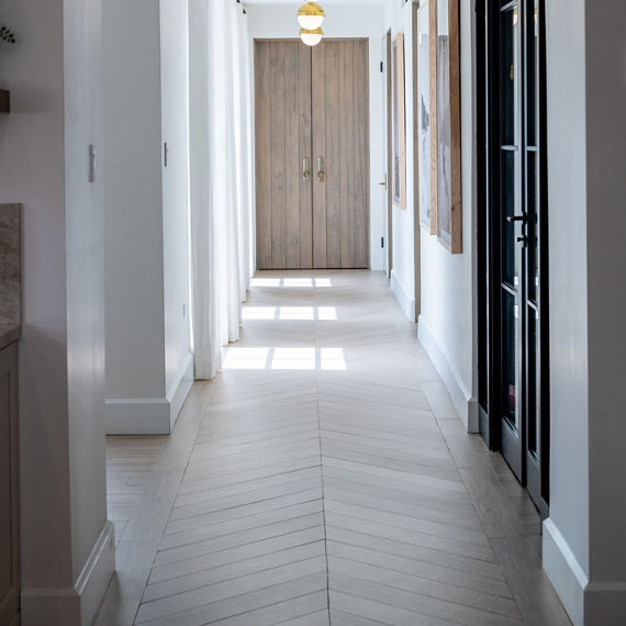 Hallway with wood look floor tile in chevron patter, white walls, and large arch at front of hallway.