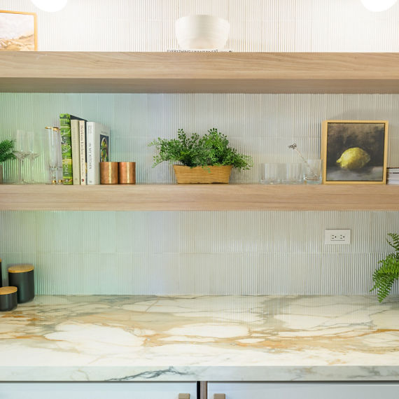 A contemporary kitchen counter with a marble-style surface is shown beneath two wooden floating shelves. The shelves display potted green plants, glassware, books, and small decorative objects, including framed art and a ceramic mug. The light-colore