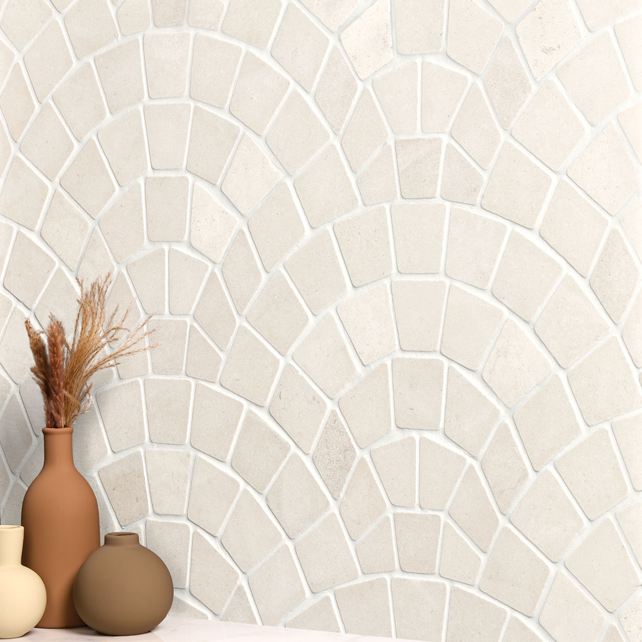A decorative wall features a light neutral mosaic tile pattern arranged in curved, fan-like shapes. In the foreground, three matte ceramic vases in earthy tones sit on a surface, one holding dried grasses. The tiles have subtle variations in beige an