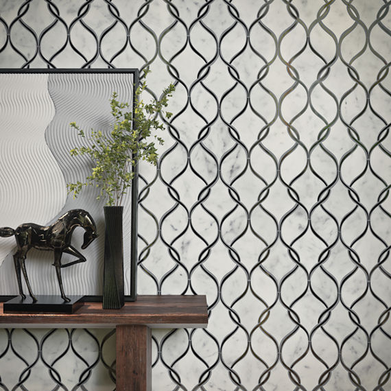 A wooden console table stands in front of a decorative marble wall with an interlocking curved pattern. On the table, a dark sculptural horse and a tall black vase with green foliage create a contemporary vignette. A textured, wavy panel in neutral t