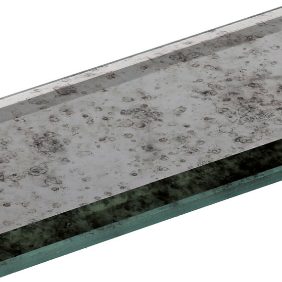 A rectangular piece of glass with an antique mirror finish is shown on a white background. The tile has beveled edges and a slightly greenish glass tint. Dark speckled oxidation patterns are visible across the reflective surface, giving it a distress