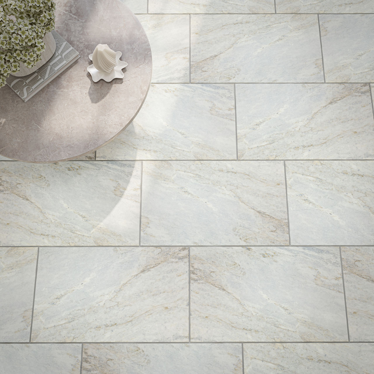 A light-colored stone tile floor is shown from above, featuring rectangular tiles with subtle beige and gray veining. In the upper left corner, a round stone table holds a decorative shell-shaped object and a leafy plant arrangement. Soft natural lig