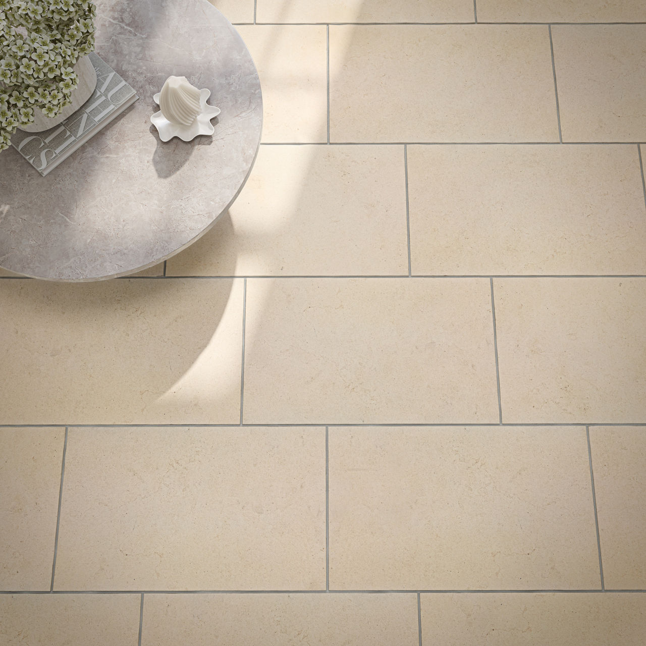 A light beige stone tile floor is shown from above, featuring a subtle speckled texture and clean grout lines. Rectangular tiles are laid in an offset pattern, creating a modern and orderly look. A round stone table with a decorative object and a pot