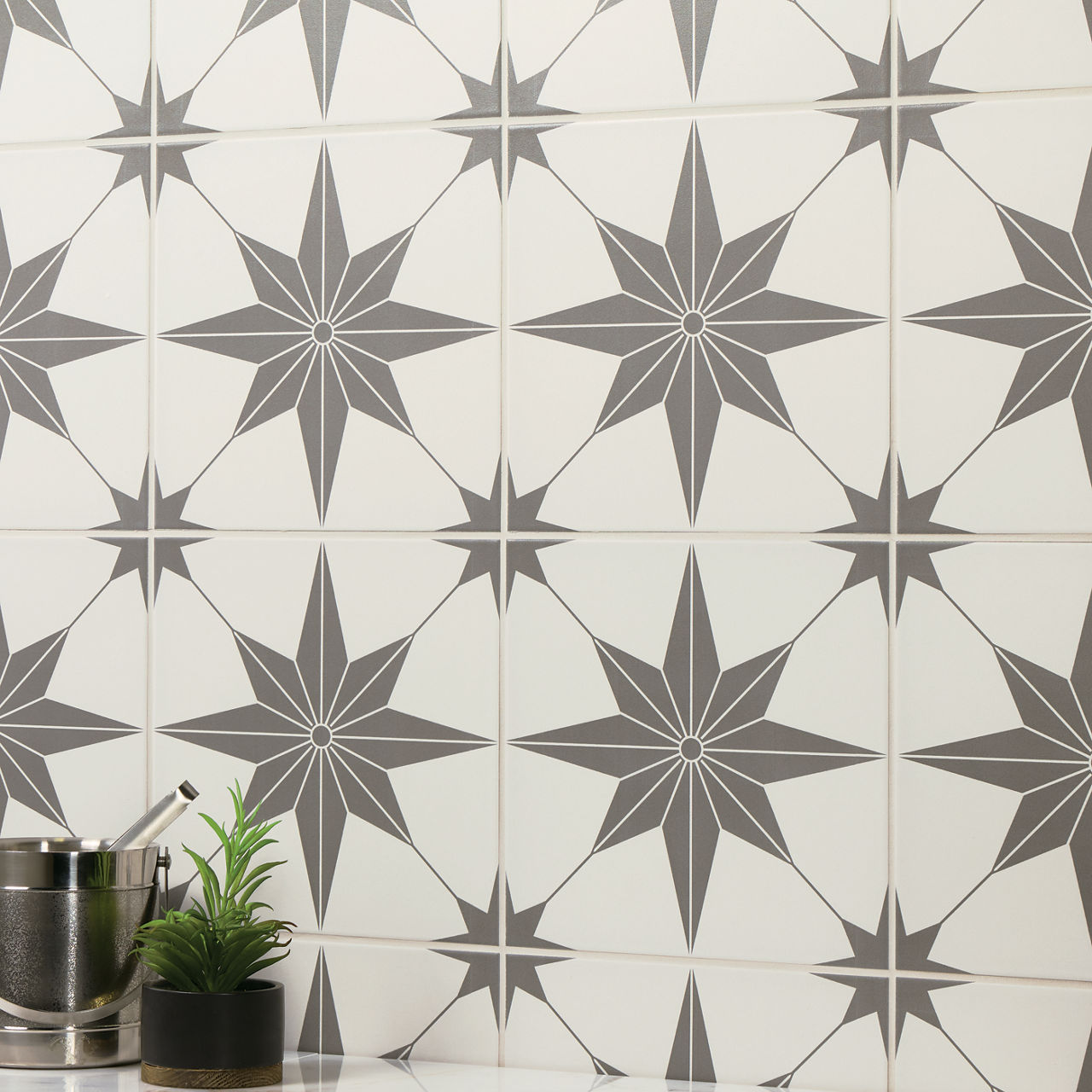 A wall is covered with ceramic tiles featuring a repeating geometric star pattern in gray on a white background. The design creates a bold, symmetrical motif that adds visual interest and rhythm. In the foreground, a small metal container with utensi