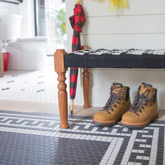 Close up of mudroom with white hexagon mosaic tile and black hexagon mosaic tile on floor in pattern, work boots, sitting bench, and foyer bathroom in background.