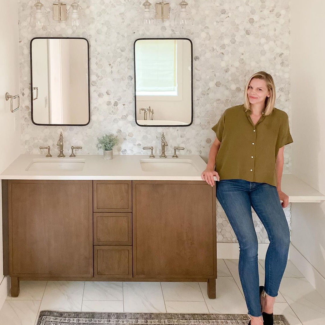 Instagram influencer Jennifer Gizzi sitting on her recently renovated bathroom vanity with white and beige marble mosaic backsplash and marble look white and gray floor tile.