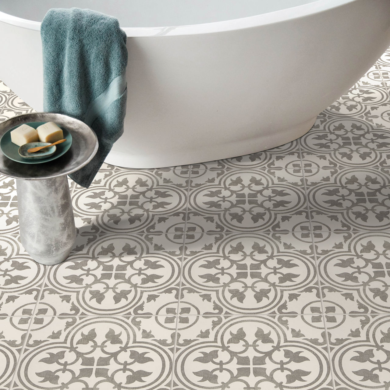 A freestanding white bathtub sits on an ornate patterned tile floor with repeating circular floral motifs. A soft teal towel hangs over the edge of the tub, adding a touch of color to the neutral space. In the foreground, a small cylindrical side tab