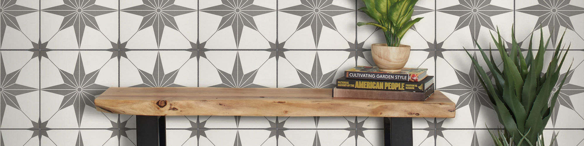 A minimalist wooden bench with black metal legs stands against a wall covered in geometric star-patterned tiles. The floor features large white tiles with subtle gray veining. A small potted plant and a stack of books sit on the bench, while a taller