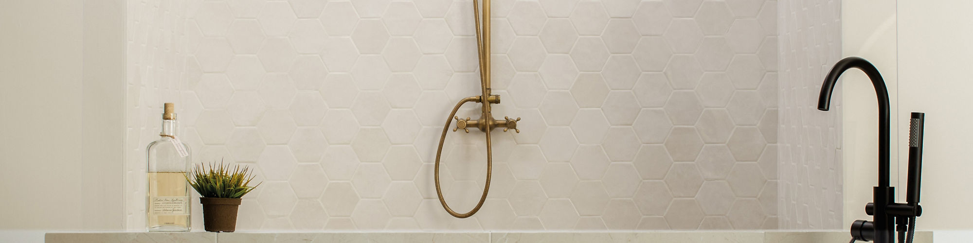 Bathroom with white hexagon tile on back wall, white curved bath tub, gold shower head, and white square floor tile.