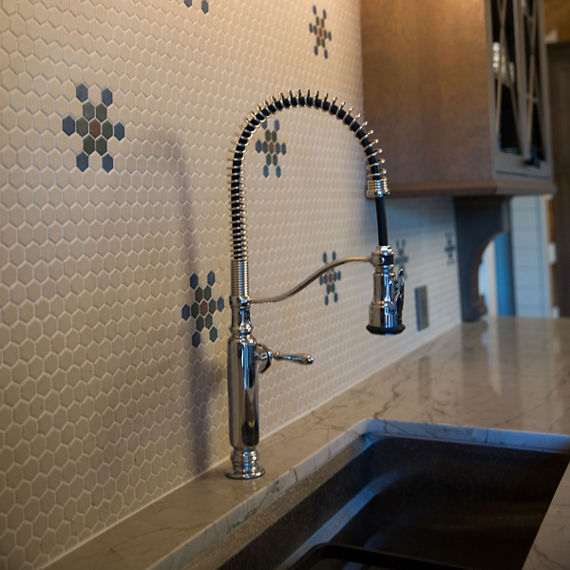 Close up of kitchen backsplash with beige hexagon mosaic tile, blue mosaic tile, green mosaic tile, on in a flower pattern, white quartz countertop, and a silver sink faucet.