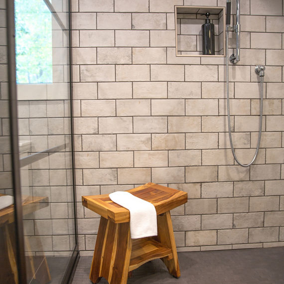 Shower with white stone look rectangle tile in a brick pattern on shower walls, small wooden stool, black hexagon mosaic tile on shower floor, and silver accents.