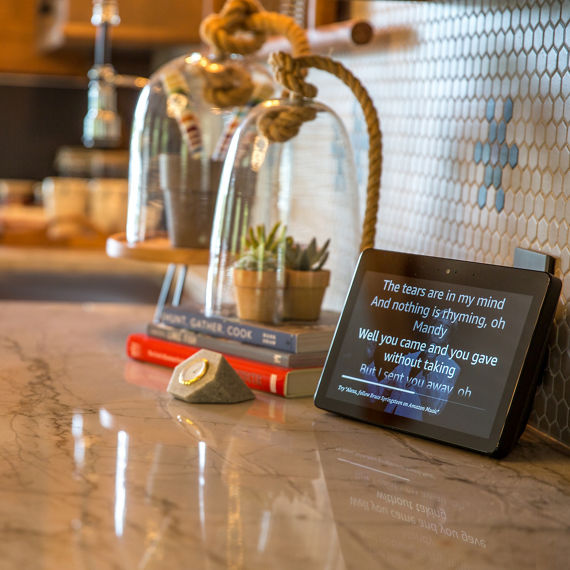 Close up of kitchen backsplash with beige hexagon mosaic tile, blue mosaic tile, green mosaic tile, on in a flower pattern, white quartz countertop, and a plant inside a glass case..