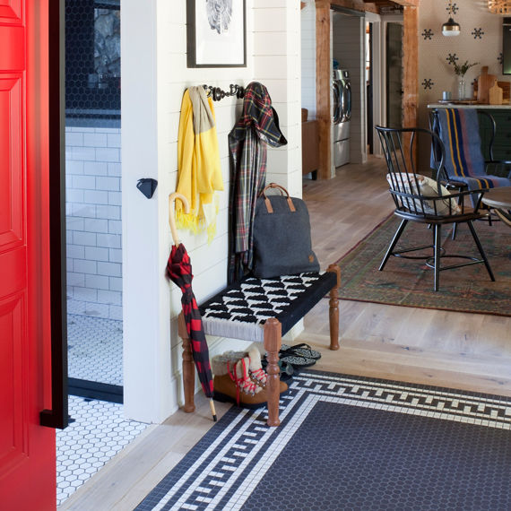Mudroom with white hexagon mosaic tile and black hexagon mosaic tile on floor in pattern, two pairs of boots, sitting bench, and kitchen in background.
