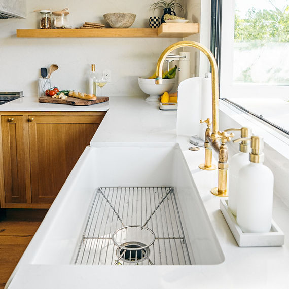 Kitchen with white marble look quartz countertop, a farmhouse sink, wooden shelves and cabinets, and a cutting board with bread and tomatoes.