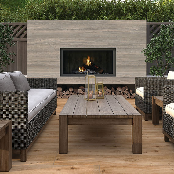Outdoor fireplace with wood look floor tile, beige marble look slab on fireplace surround, and a wooden coffee table.