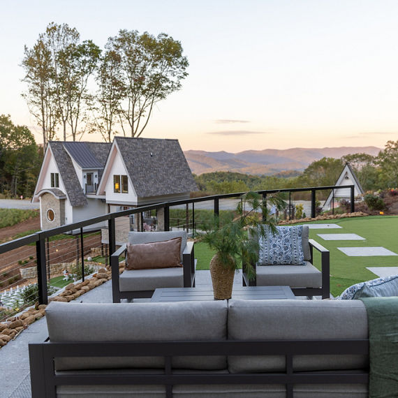 A contemporary outdoor patio features cushioned seating and a small table with a potted plant, set against a backdrop of rolling hills and distant mountains. 