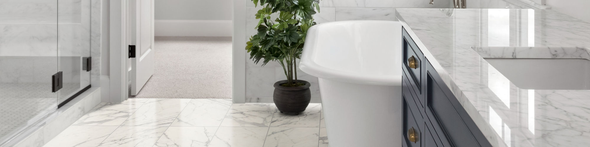 A modern, upscale bathroom features a white freestanding bathtub beside a large window. Marble tile flooring and a glass-enclosed shower create a clean, elegant look. A navy blue vanity with marble countertop and brass hardware adds contrast to the p
