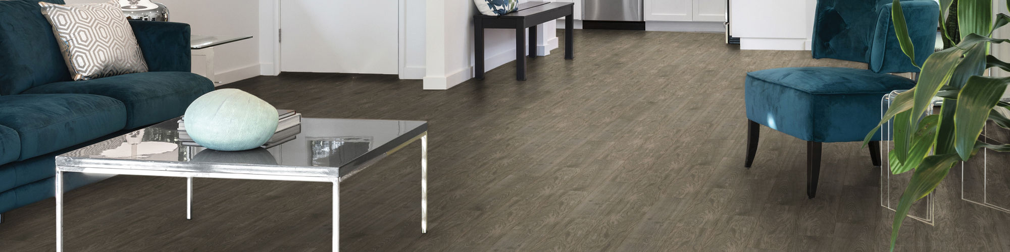 Bright open-plan living space features dark wood-look flooring and white walls. A teal sofa and matching chair sit around a glass coffee table with decorative accents. The room extends into a white kitchen with stainless steel appliances and large wi
