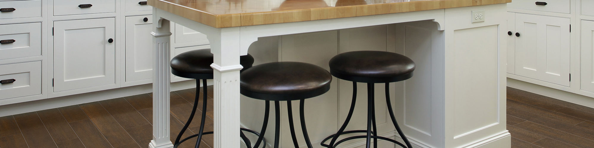 Spacious contemporary kitchen features white cabinetry and a central wooden island with three metal stools. Large windows with blinds bring in natural light, highlighting the dark wood flooring and black countertops. Hanging pot rack, pendant lights,