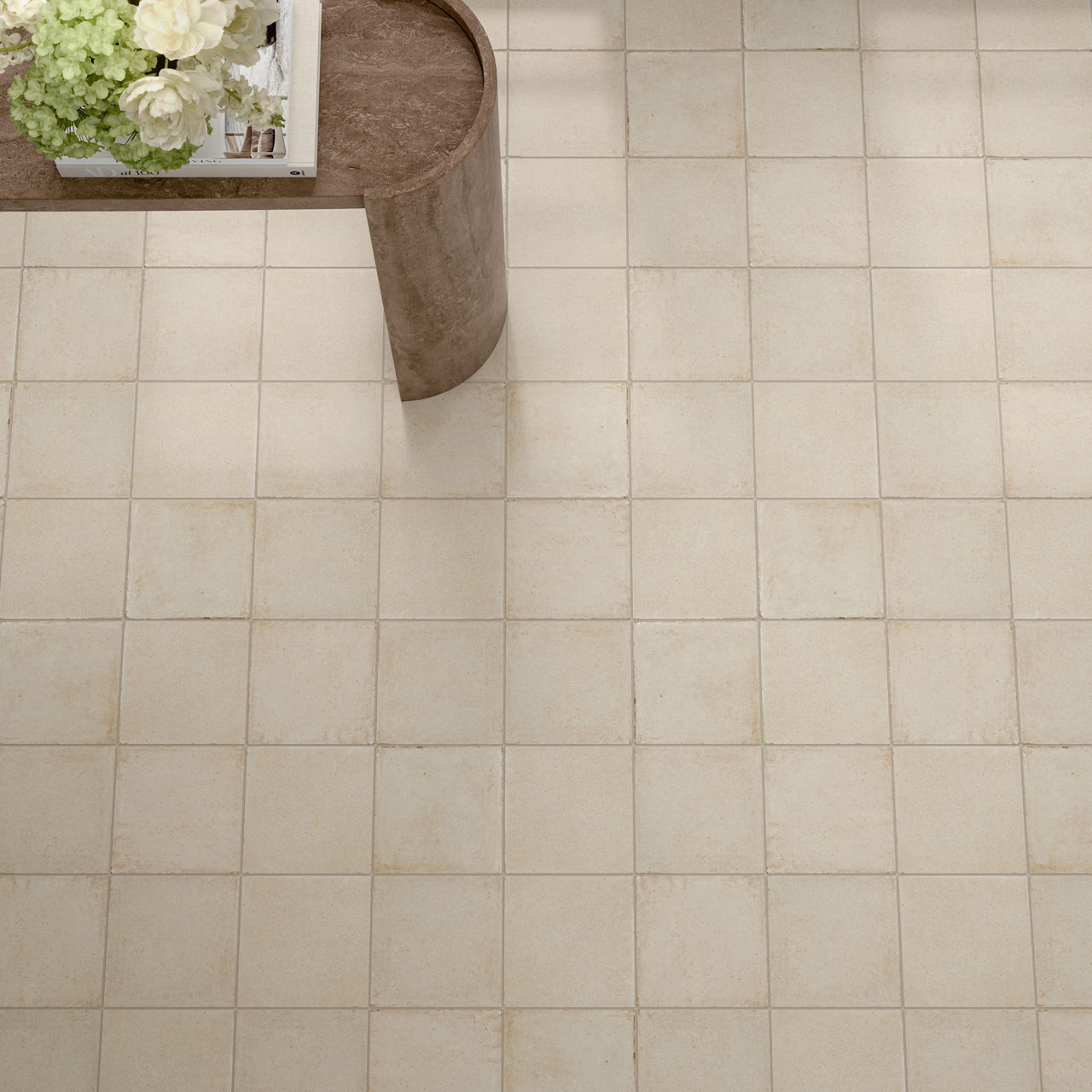 A beige tiled floor is seen from above, showing a grid of evenly spaced square tiles. In the upper left corner, part of a curved wooden or stone table is visible. On the table sits a white tray holding white and green flowers. The overall scene feels