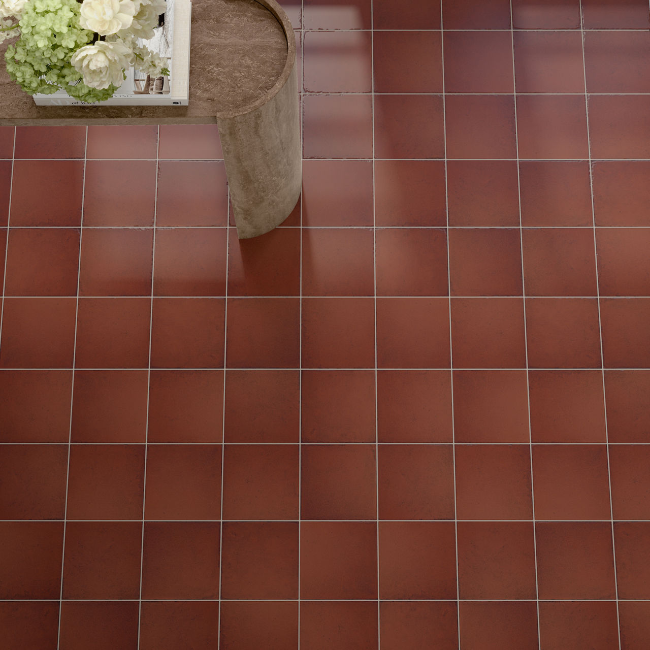 Overhead view of a red ceramic tiled floor with evenly spaced grout lines. In the upper left corner, a small table or stand holds a white tray filled with light green and white flowers. A cylindrical stone or concrete pedestal sits beside the tray, a
