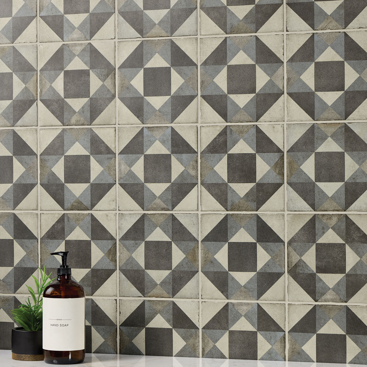 A wall covered in square ceramic tiles features a bold geometric diamond pattern in gray and off-white tones. In the lower left corner, a brown pump bottle labeled "HAND SOAP" stands on a surface beside a small green plant in a black pot. The scene s