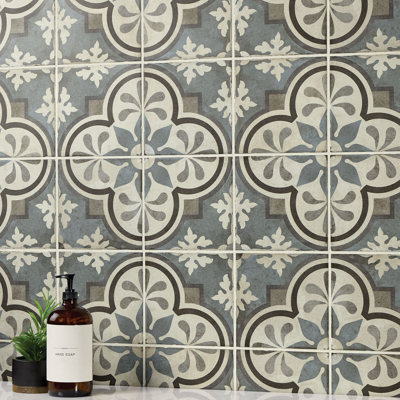 A wall covered in ornate patterned tiles features repeating floral and geometric motifs in muted blue, gray, and cream tones. The design creates a vintage, Mediterranean-inspired backdrop with a slightly weathered look. In the foreground, a brown gla