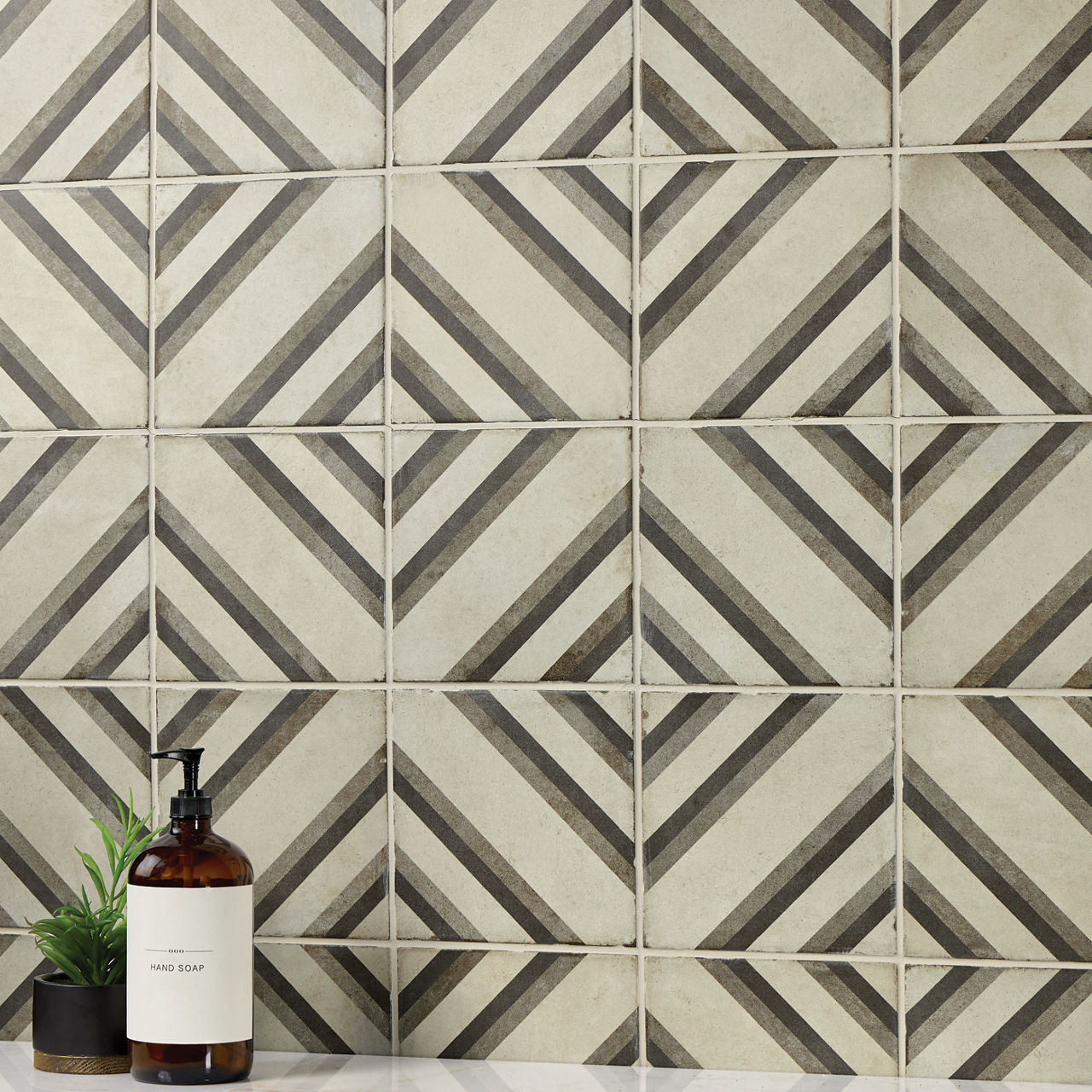 A geometric tiled wall with diagonal striped patterns forms the backdrop of this scene. The tiles create a repeating diamond motif in neutral beige and gray tones. In the foreground, a brown glass hand soap bottle with a pump sits on a countertop bes