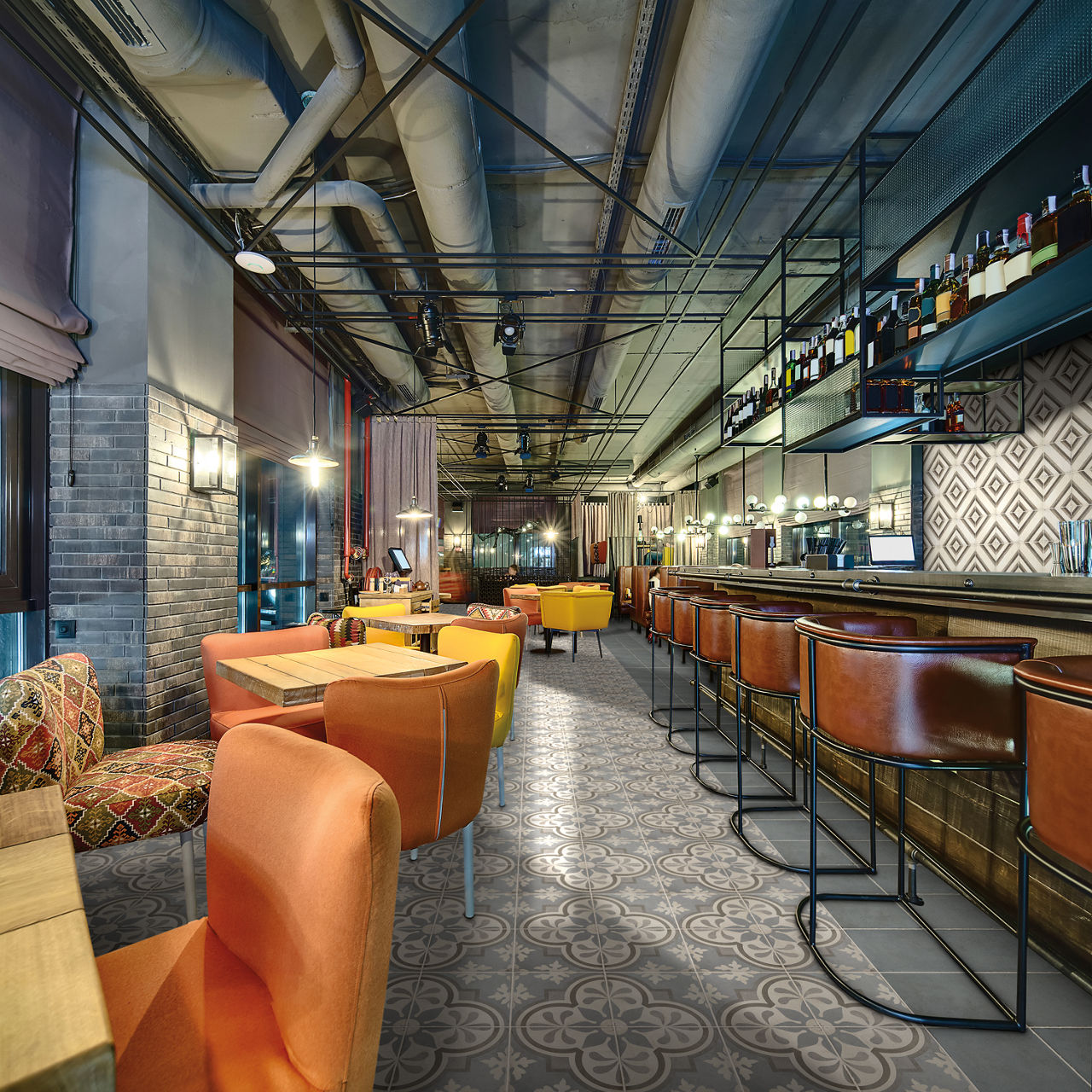 A contemporary bar interior features a long counter with high leather stools and a patterned tile floor. Colorful armchairs and small tables create a cozy lounge area in the foreground. Overhead, exposed ductwork and industrial lighting contribute to
