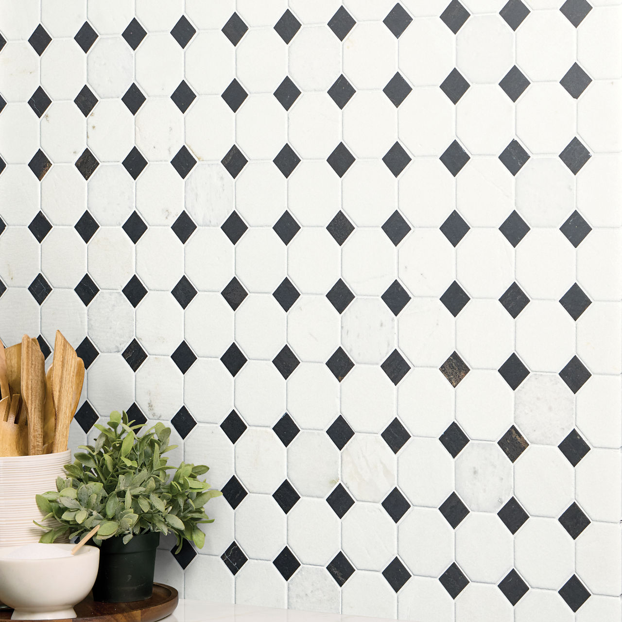 A white octagon tile wall with small black diamond insets forms a geometric backdrop. The pattern creates a clean, modern look with strong contrast between light and dark tiles. In the foreground, a potted green plant and wooden utensils in a contain