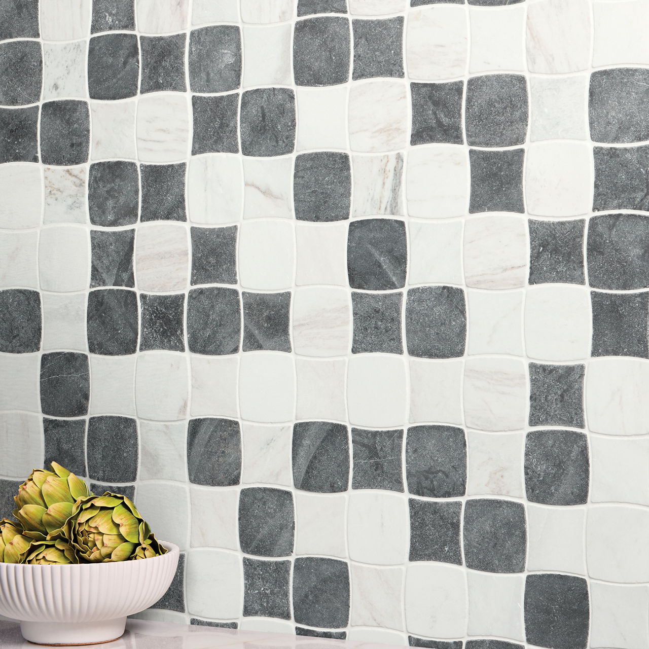 Curved square tiles in alternating gray and white create a checkered wall pattern. The surface has a subtle mix of matte and marble-like textures, giving the design a contemporary look. In the foreground, a white ribbed bowl holds fresh artichokes on