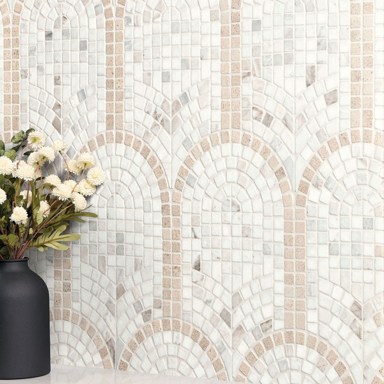 A decorative mosaic tile wall features an arched pattern in soft beige and white tones. The tiles are arranged in a repeating geometric design that creates a subtle architectural feel. In the foreground, a matte black vase holds white and yellow flow