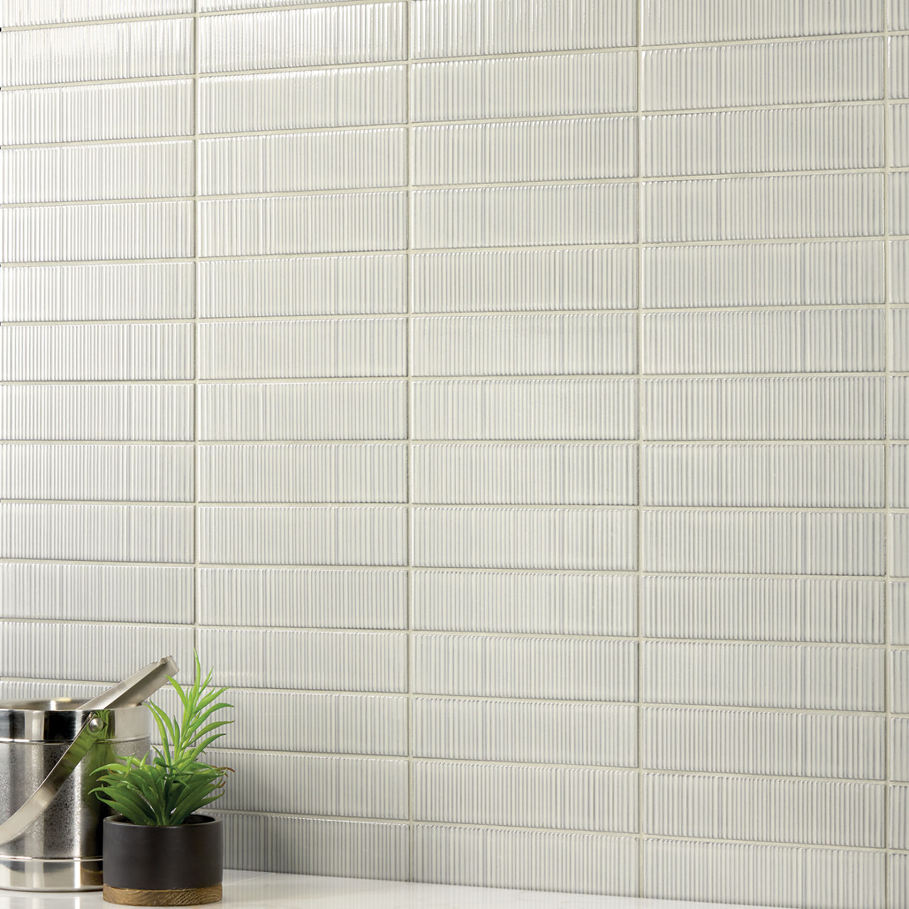 White rectangle wall tile with ripple design, silver bucket, and small plant on side.