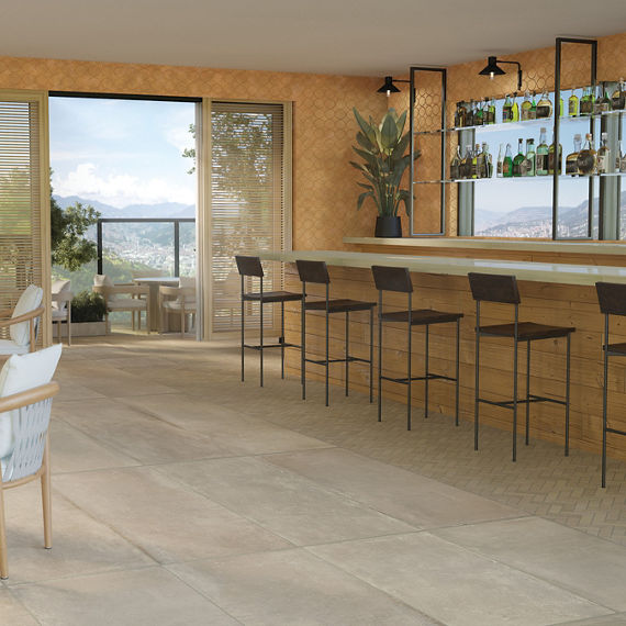 Restaurant with a mountain view, beige large format floor tile and matching mosaic that look like concrete, woodgrain bar with bar stools, table and chair with white cushions.