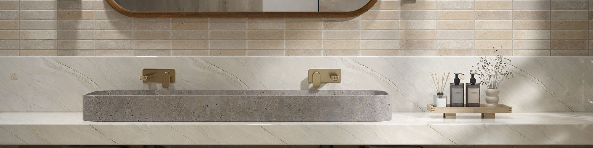 A contemporary bathroom features a long stone double sink set into a sleek floating countertop. The wall behind the vanity is covered in warm neutral tiles, complemented by an oval mirror and two vertical wall sconces. Woven storage baskets with towe