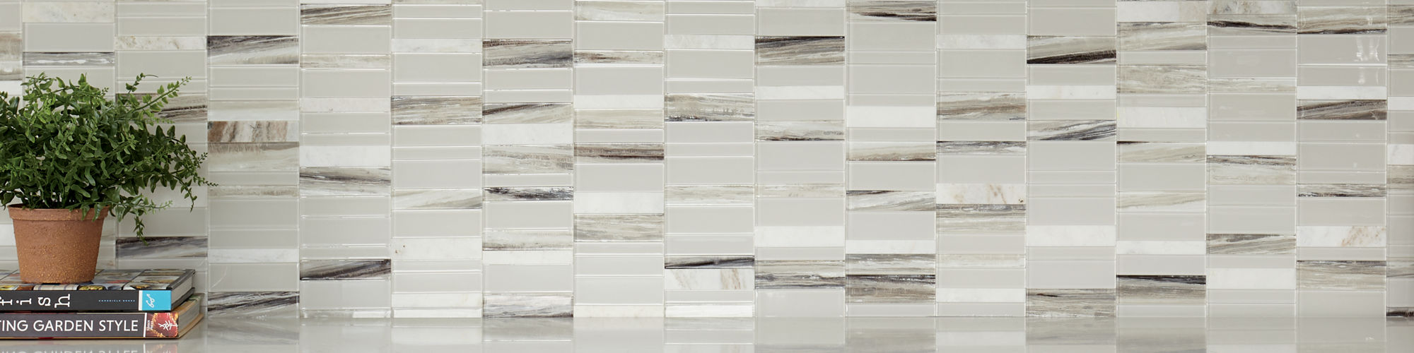 Kitchen with white natural stone rectangle mosaic on backsplash, white cabinets, white countertop, stack of books, and pot with greenery on stack of books.