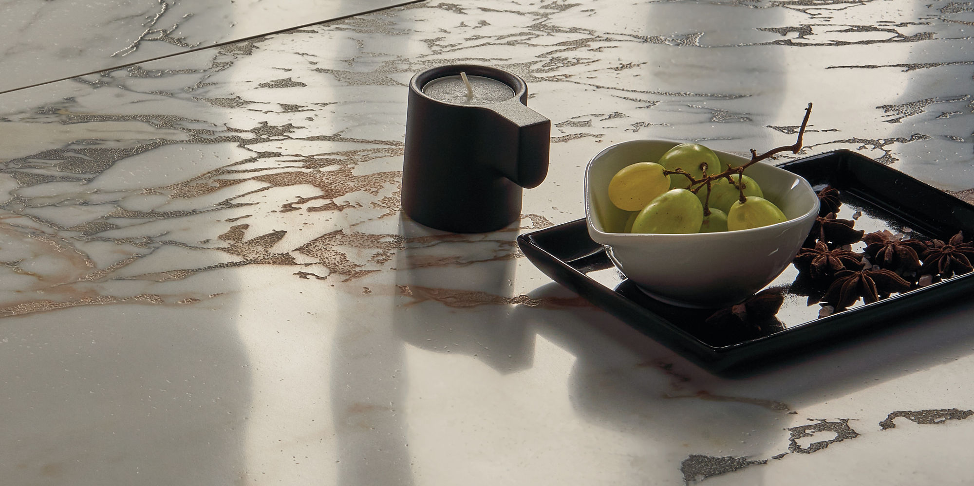A modern marble surface is adorned with a small ceramic mug and a black tray holding a bowl of green grapes. Soft sunlight casts gentle shadows across the scene, highlighting the elegant veining of the marble. The overall mood is calm and minimalist,