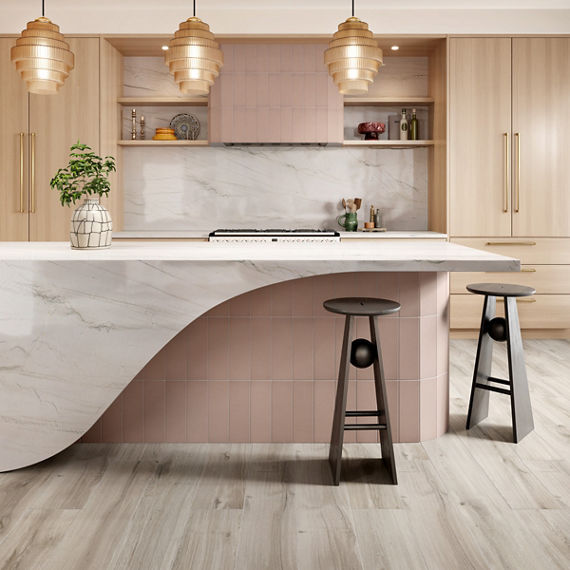 Kitchen with pink wall tile with linear pattern on island counter and stove vent, white marble look slab on island countertop and stove backsplash, light wood cabinets, and light wood look floor tile.