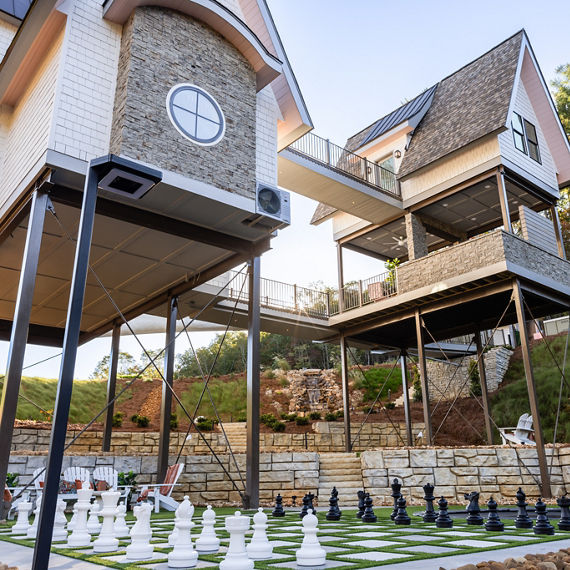 Two contemporary houses elevated on stilts and connected by a bridge on a sloped landscape, with a large paver tile chessboard featuring oversized pieces in the foreground, surrounded by greenery and stone retaining walls in clear daylight.