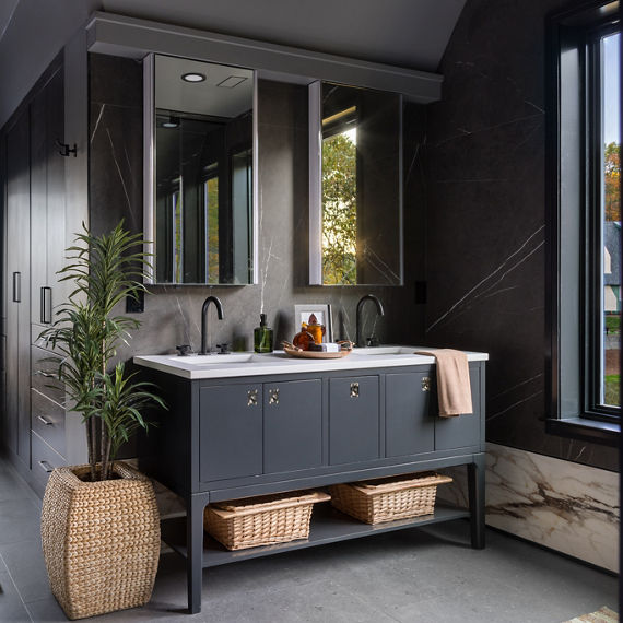 Bathroom with a double?sink vanity featuring dark cabinetry and gold hardware, accented by a woven basket planter, under?sink storage baskets, and a large mirror, with natural light from a window and a glimpse into the adjoining room.