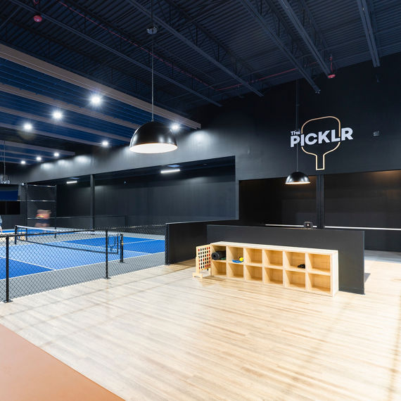 indoor pickleball court with light wood vinyl floor tile, blue courts, and black walls.