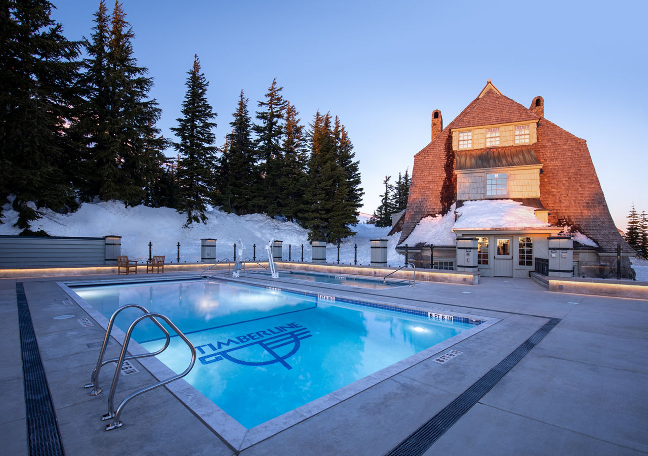 A heated outdoor swimming pool is featured in front of a rustic mountain lodge, surrounded by snow-covered trees and landscape.