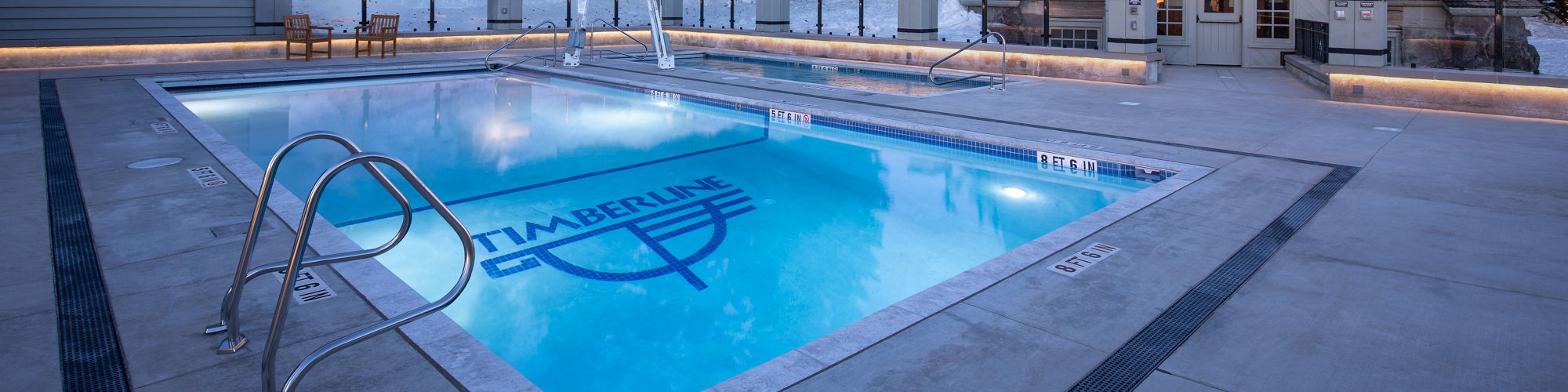 A heated outdoor swimming pool is featured in front of a rustic mountain lodge, surrounded by snow-covered trees and landscape. 