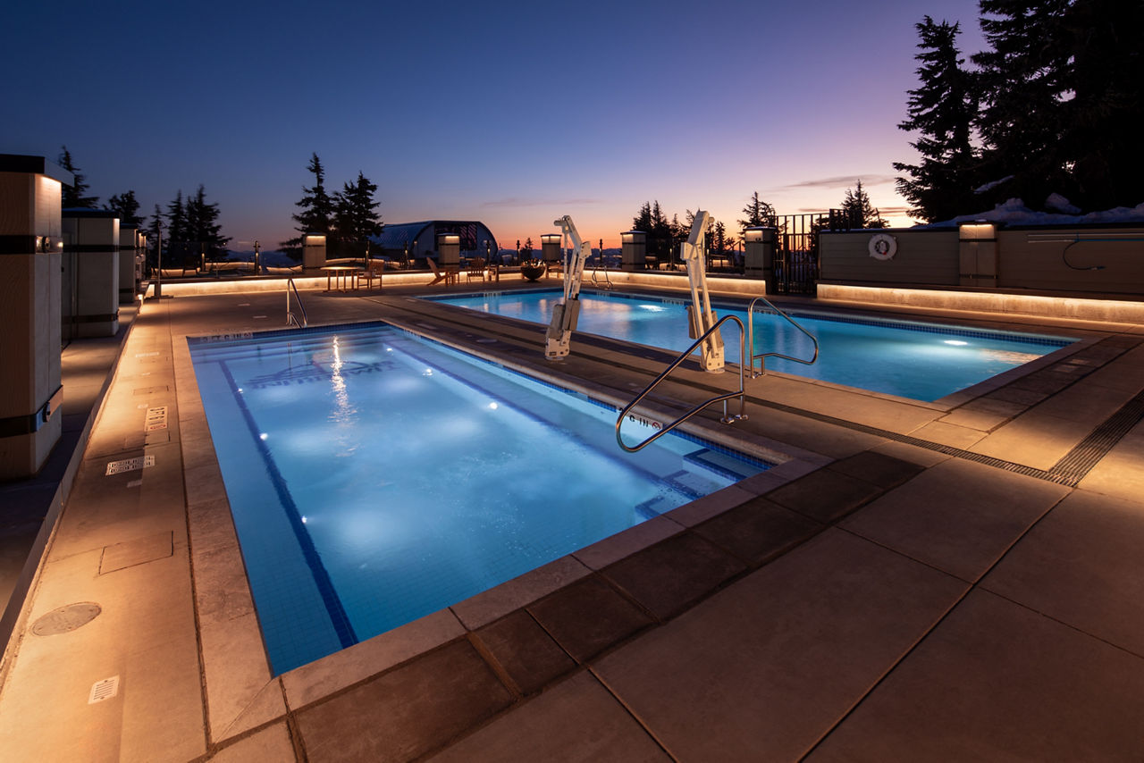 Two rectangular swimming pools are featured in an outdoor setting at dusk, surrounded by modern railings and deck. 